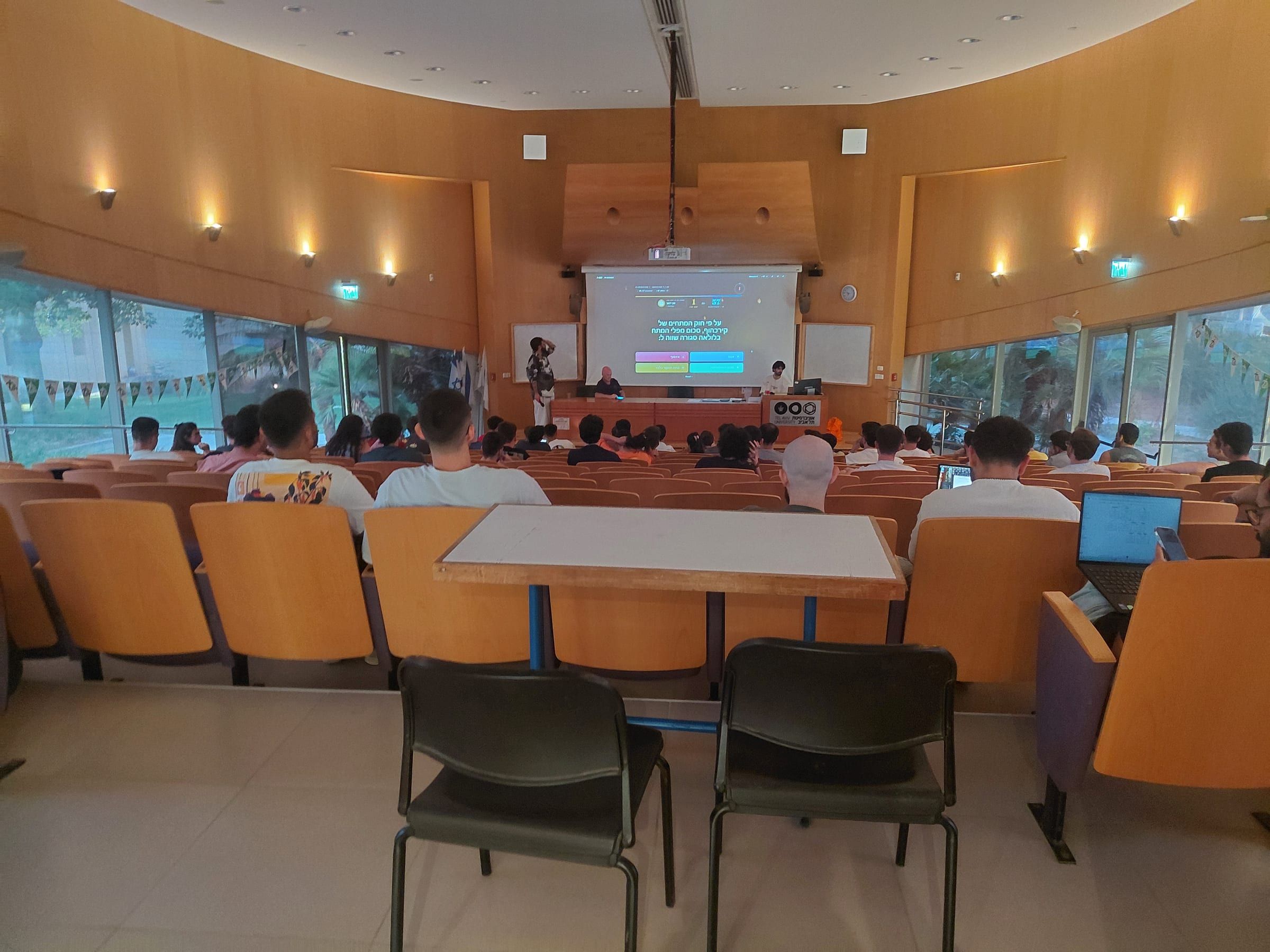 A Tel Aviv University lecture hall seen from the back, most seats taken, students facing a projected 1 vs 100 question in Hebrew with four coloured answer options and a countdown.