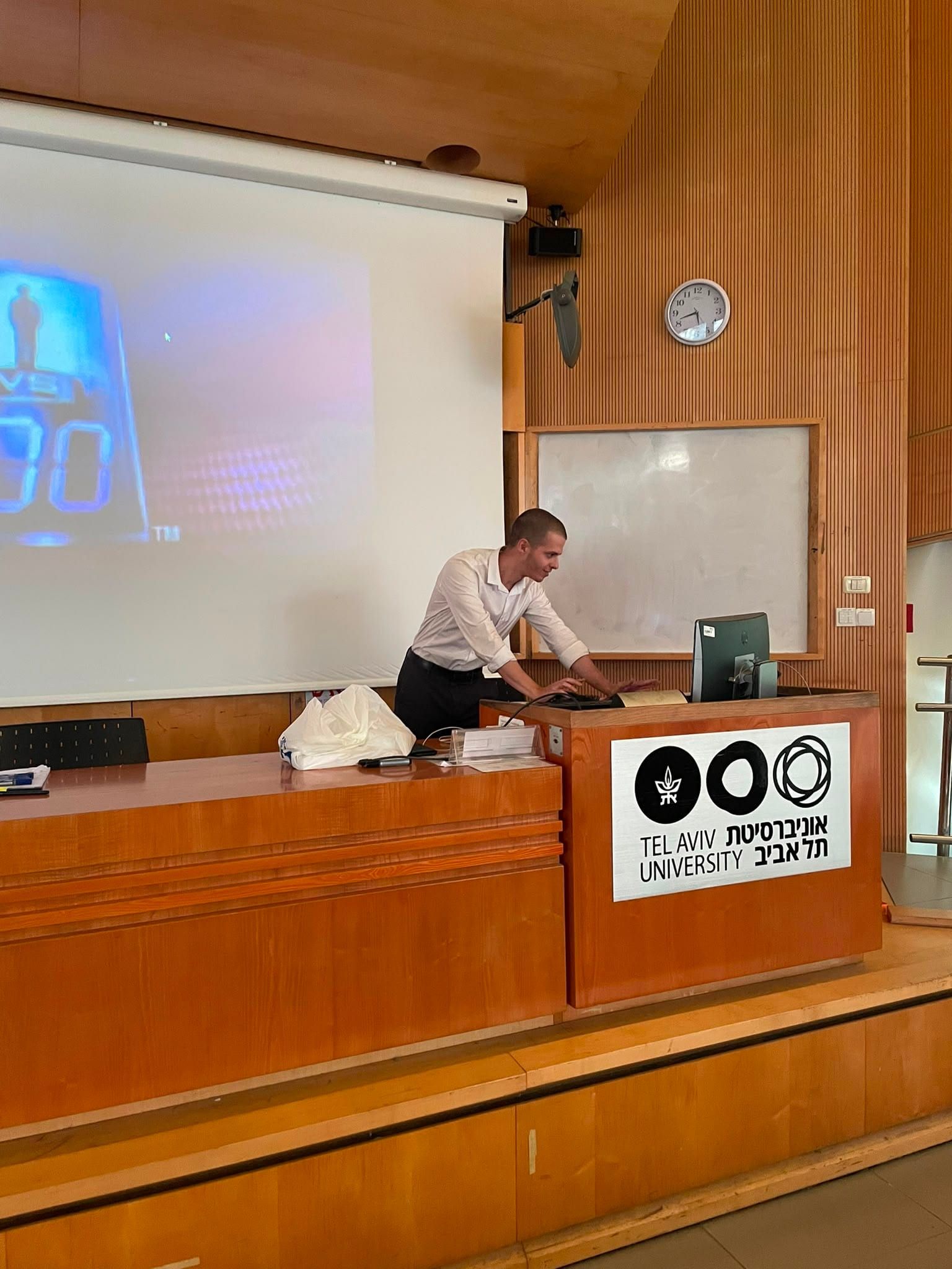 David Mashiah at the lecture-hall lectern working at the podium computer, the Tel Aviv University logo on the panel beside him and the 1 vs 100 title card projected behind.