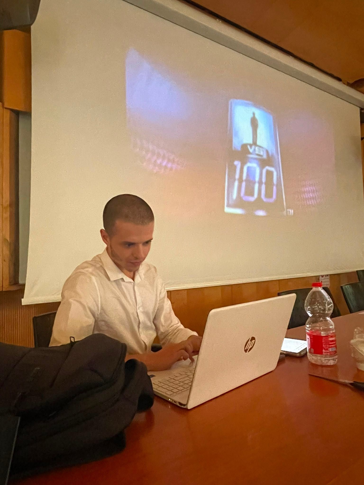 David Mashiah seated at a table in the lecture hall working on a laptop, with the 1 vs 100 title card projected on the screen behind him.